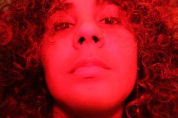 Imagem com filtro vermelho mostra o rosto de uma mulher de cabelos encaracolados, expressão séria, encarando a câmera. Ela tem piercing de argola no nariz. 