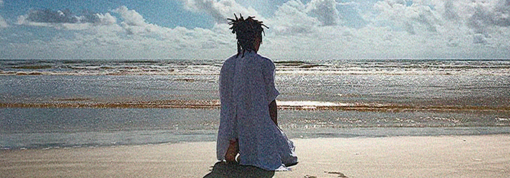 Uma mulher está sentada, de joelhos, em uma praia, de costas para a câmera, de frente para o mar. Ela usa roupas brancas. 